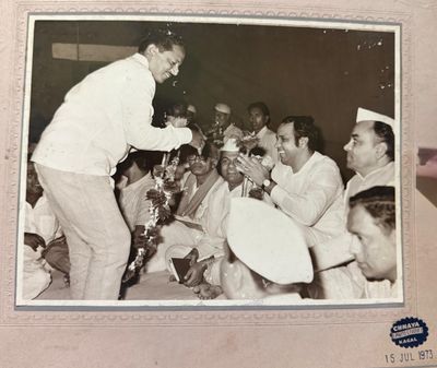 A felicitation ceremony — being garlanded and honoured amid a seated crowd.